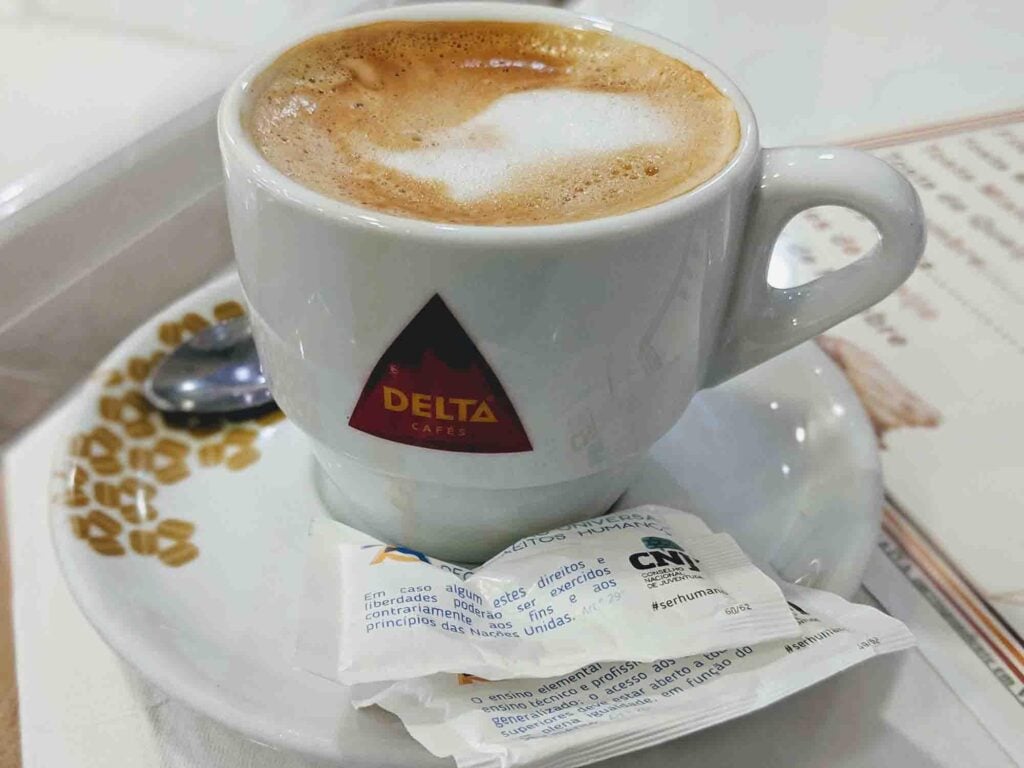 A Meia de Leite cup of coffee in a white mug with the "Delta Cafés" logo on it, placed on a saucer with a small spoon on the side. There are two sugar packets on the saucer beside the cup. The background shows part of a menu with text, evoking the cozy ambiance of a café in Portugal.
