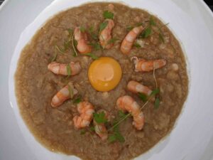 A white plate contains a creamy beige dish garnished with several prawns arranged in a circle. In the center, there is a raw egg yolk. Fresh green herbs are sprinkled throughout the dish, adding a touch of color.