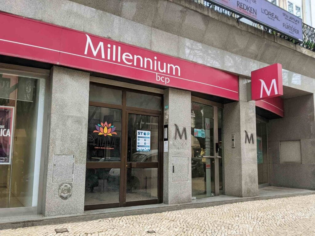 Photograph of the entrance to a Millennium bcp bank branch. The facade features a prominent red and white sign with the bank's logo above the doorway. The building exterior is made of stone, and there's a plaque next to the door. Cobbled stone pavement is in front.