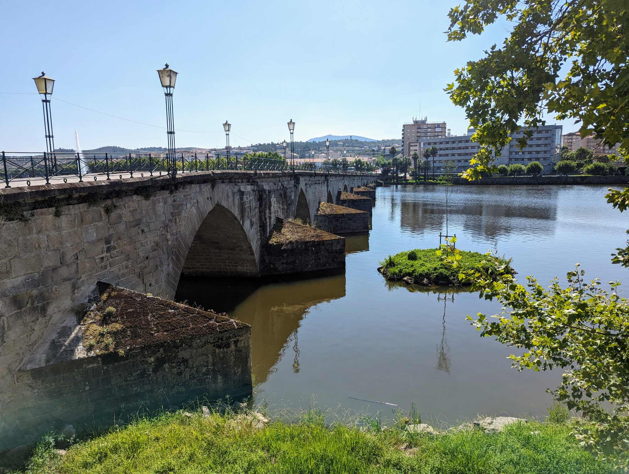 A historic stone bridge with arched supports stretches across a calm river, evoking the charm of Portugal. Street lamps line the bridge, and buildings can be seen in the background. A small, grassy island with a tree adds to the scenic view, while trees frame the right side.