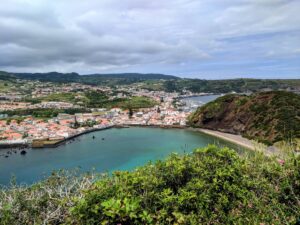 A picturesque coastal town in Faial with white and red-roofed buildings nestled along a curved shoreline. Clear turquoise water gently laps against the beach, and lush greenery covers the surrounding hills. The sky is partially cloudy, casting soft light over the scene.