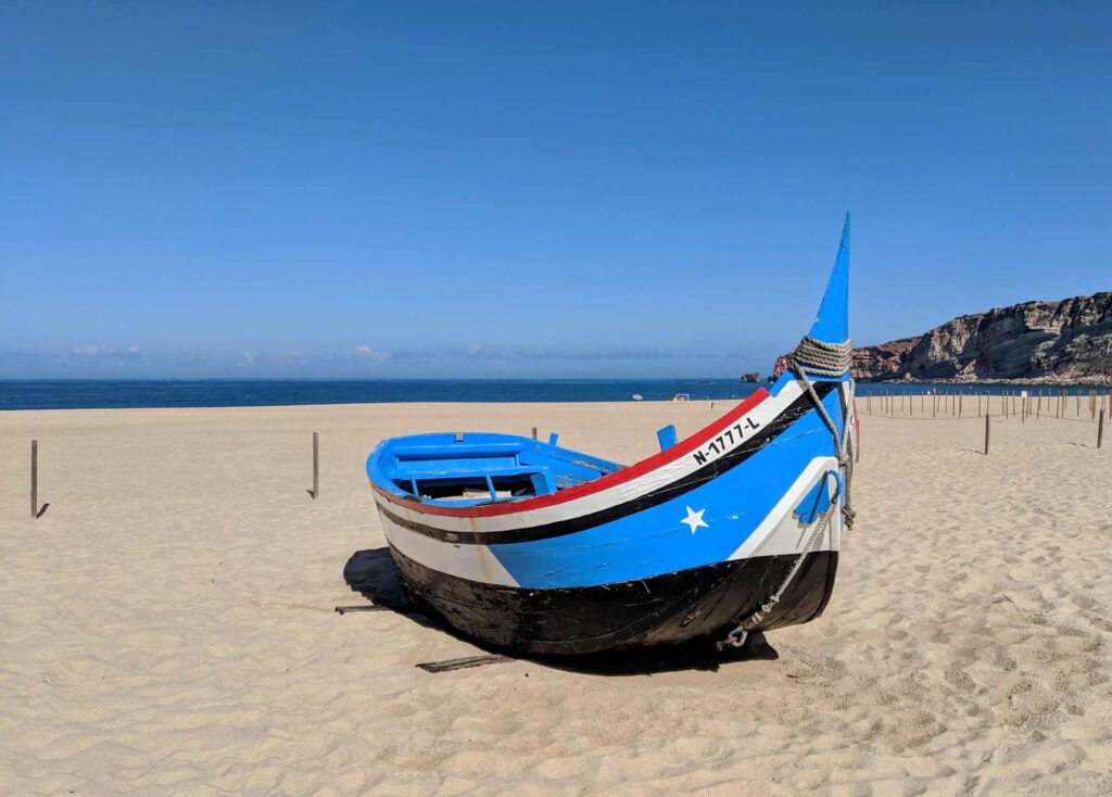A small, colorful fishing boat is beached on a sandy shore under a clear blue sky in picturesque Portugal. The boat is painted in vibrant blue, white, and red, with a blue anchor in the bow. The calm ocean and rocky cliffs are visible in the background.