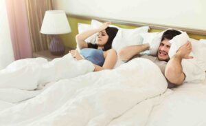A man and a woman lying in bed under white blankets appear disturbed. The man, on the right, covers his ears with a pillow, while the woman, on the left, looks annoyed and is holding her head. A lamp on a nightstand is in the background.