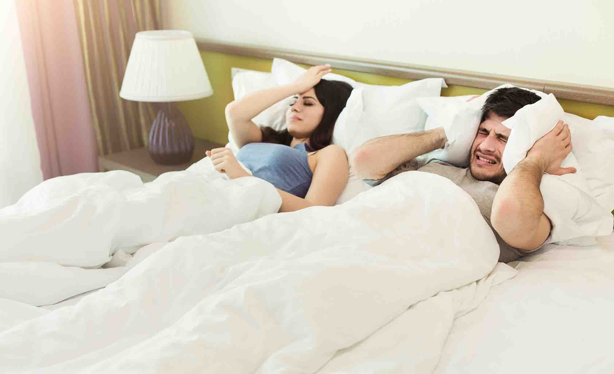 A man and a woman lying in bed under white blankets appear disturbed. The man, on the right, covers his ears with a pillow, while the woman, on the left, looks annoyed and is holding her head. A lamp on a nightstand is in the background.