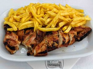 A white plate filled with grilled chicken pieces and a large serving of golden, crispy french fries. The plate is placed on a white napkin that has a logo and the word "Churrasqueira" on it, evoking the flavors of Portugal.