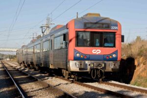A blue and red Commuter train labeled "2295" rides on a double-track railway under clear skies. The train, belonging to Comboios de Portugal (CP), is moving through a rural area with power lines and a bridge visible in the background, showcasing the scenic beauty of Portugal.