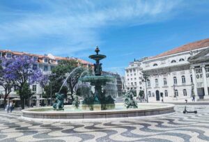A picturesque public square in Portugal with a large ornate fountain featuring statues, surrounded by historic buildings under a clear blue sky. Purple jacaranda trees are in bloom, and pedestrians walk leisurely on the intricate black and white cobblestone pattern.