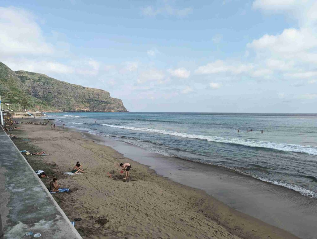 A sandy beach in Portugal on a partly cloudy day with gentle waves washing ashore. People are sunbathing, playing, and swimming in the sea. There's a concrete wall on the left side with hills and vegetation in the background. The scene is serene and relaxed.
