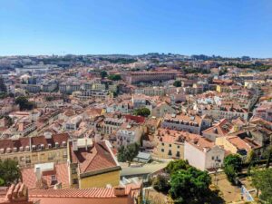 A bright and clear day view of a sprawling cityscape in Lisbon, Portugal, featuring numerous tightly packed buildings with red-tiled roofs. The city is set against a backdrop of rolling hills, with many clusters of buildings extending into the distance under a clear blue sky.
