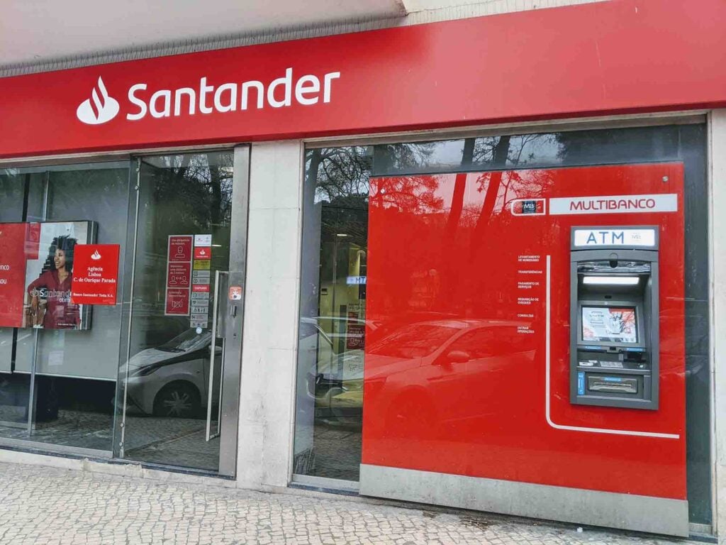Santander bank in Portugal with red signs, glass front, and a Multibanco ATM on the right; trees and cars reflect in the windows.