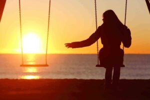 A silhouette of a person sitting on a swing by the beach at sunset, with one arm outstretched towards the empty swing beside them. The sky is a vibrant orange, and the sun is setting over the ocean in the background.