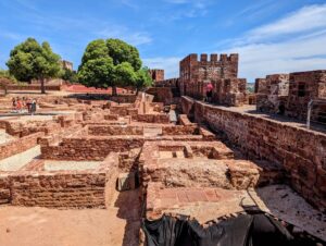 A historic stone fortress with intricately patterned walls and multiple levels, surrounded by green trees under a clear blue sky. Visitors are exploring the site, and the ancient ruins are spread across the foreground.