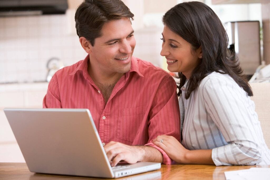 A smiling couple sits at a table looking at a laptop. The man in a red shirt types on the keyboard while the woman in a white striped shirt leans on his shoulder, both appearing engaged and happy. They are in a bright, cozy kitchen environment.