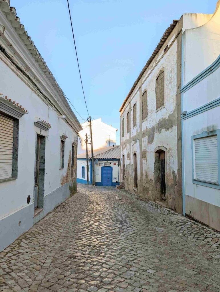 A narrow cobblestone street lined with whitewashed buildings in varying states of disrepair. The buildings have shuttered windows, and one at the end of the street features a blue door. A bright blue sky is visible overhead.