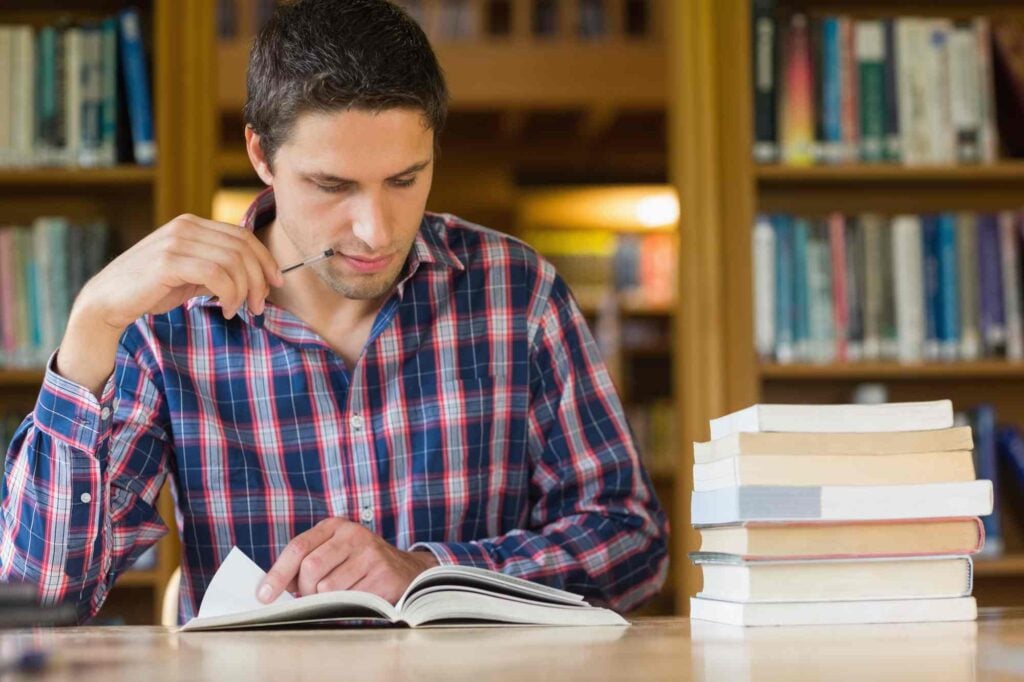 A man in a plaid shirt sits in a library, reading a book and holding a pencil to his mouth. He is surrounded by shelves filled with books, with a tall stack of books beside him on the table. The atmosphere is studious and quiet.
