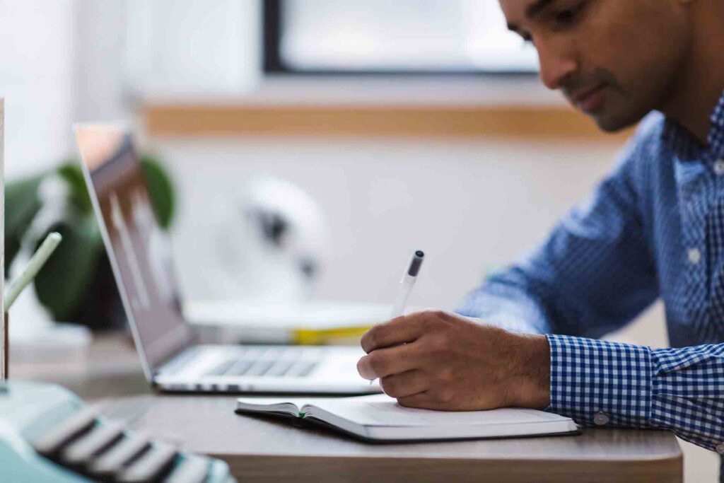 A man in a blue checkered shirt is sitting at a desk, writing in a notebook with a pen. In front of him is an open laptop, and there's a blurred background with a window, some indoor plants, and office items.