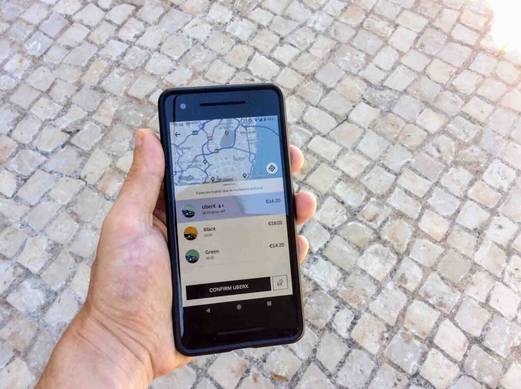 A hand holding a smartphone displaying a ride-hailing app, showing various car options including UberX and Black, with estimated fares in Euros. The background consists of a cobblestone pavement.