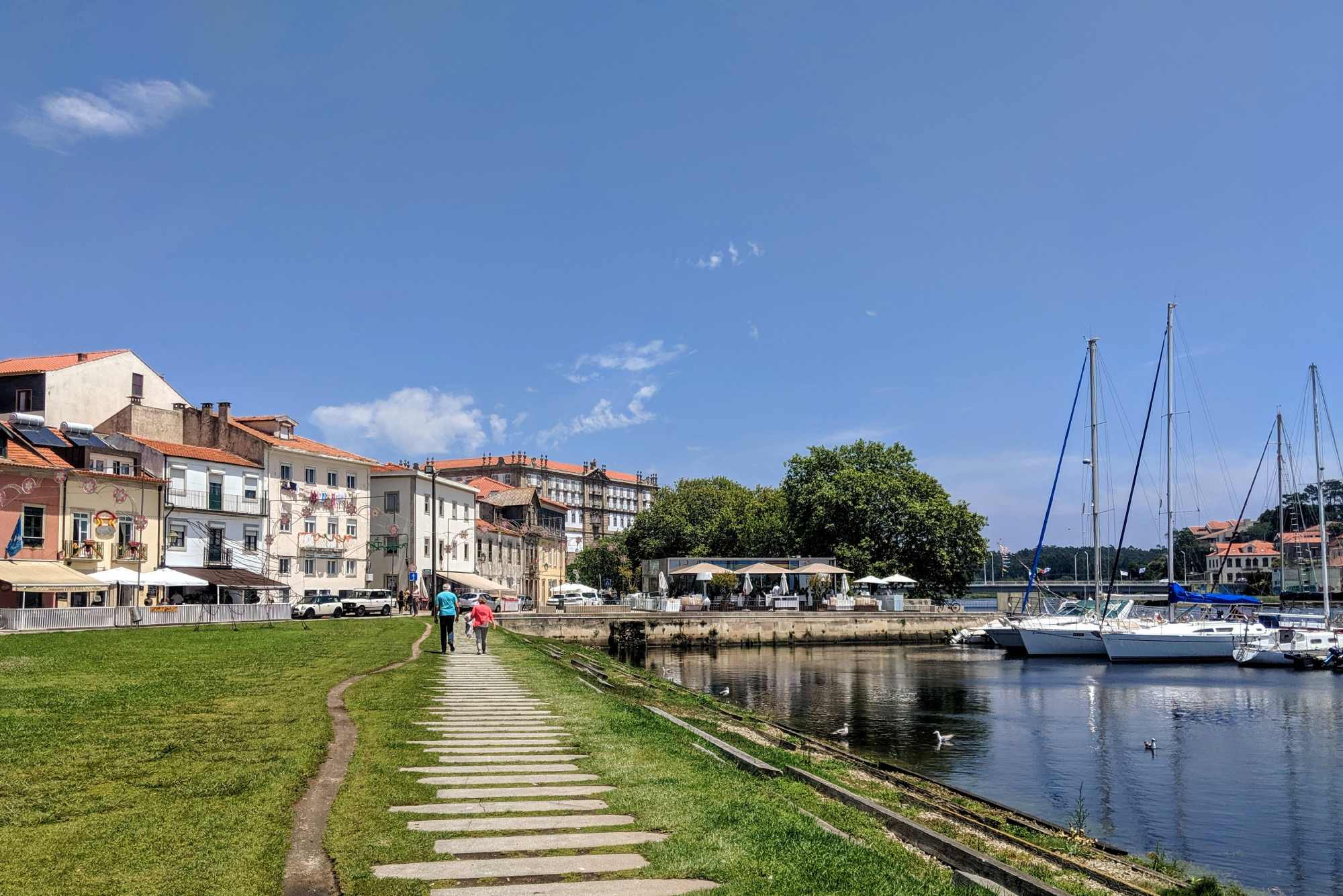 A picturesque riverside walkway in Portugal with people strolling along stepping stones on the grass. The path is flanked by charming buildings on the left and a marina with sailboats on the right. The sky is clear and blue, adding to the serene atmosphere.
